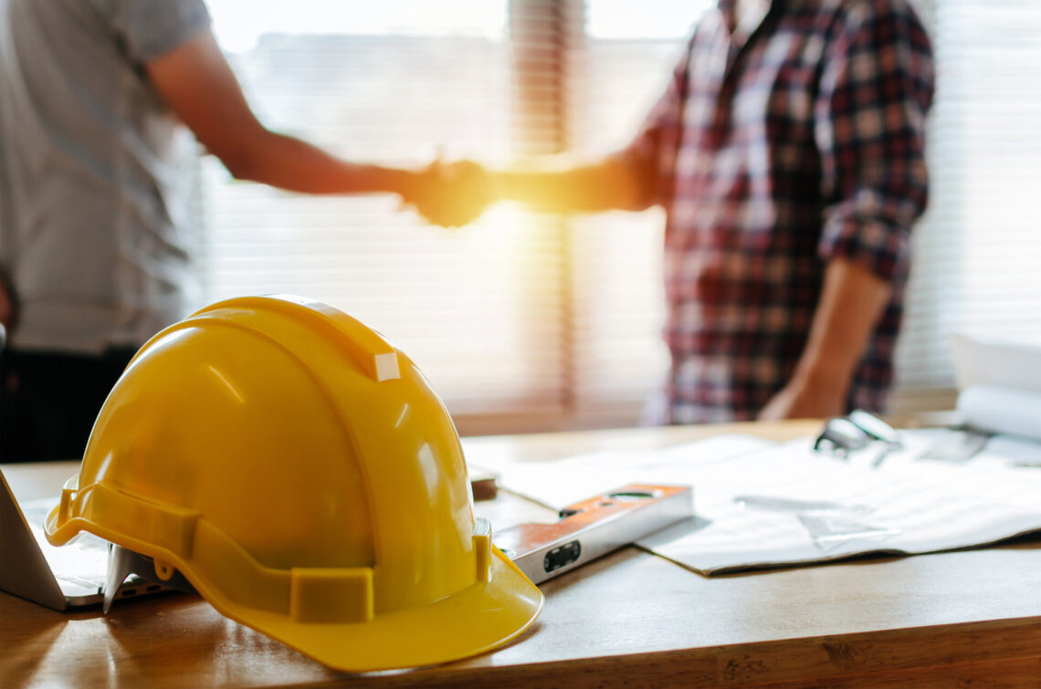 gelber Schutzhelm auf Arbeitsplatz Schreibtisch mit Bauarbeiter Team Hände schütteln Begrüßung Start-up-Plan neues Projekt Vertrag im Büro-Center auf der Baustelle