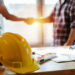 gelber Schutzhelm auf Arbeitsplatz Schreibtisch mit Bauarbeiter Team Hände schütteln Begrüßung Start-up-Plan neues Projekt Vertrag im Büro-Center auf der Baustelle